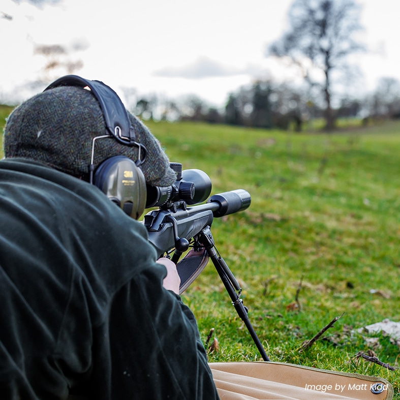 A deer stalking lying down aiming through the scope of their rifle