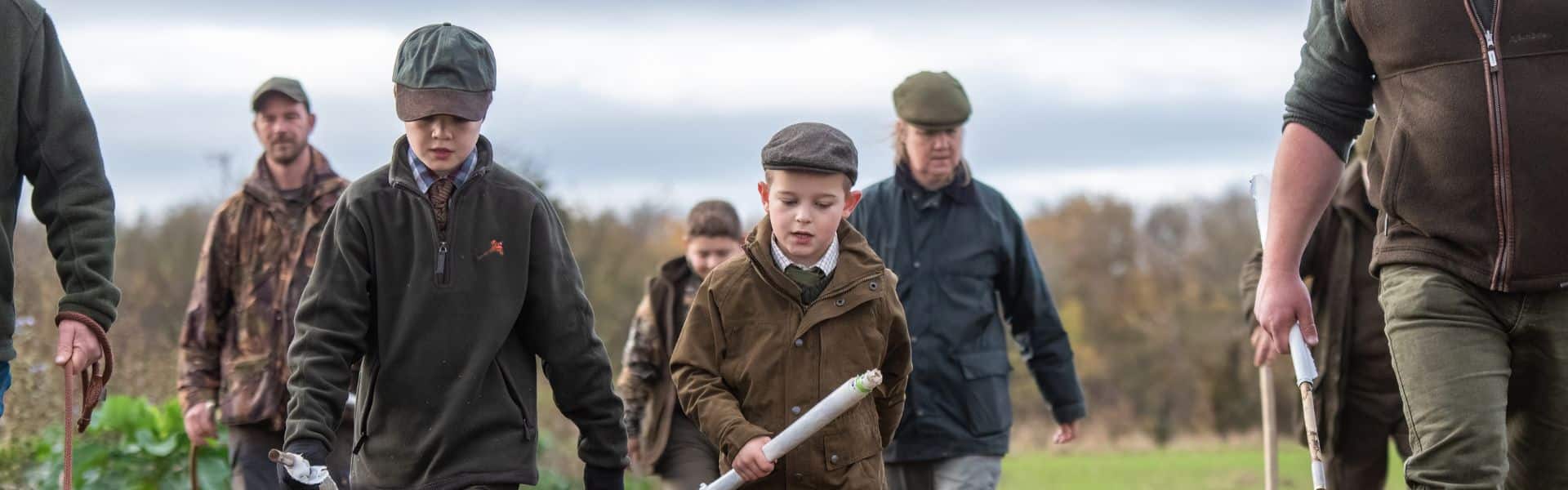 A group of game shooters and two children walking through a field holding beater flags