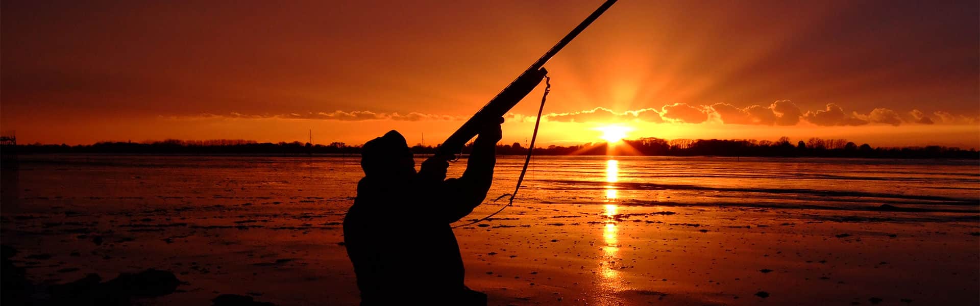 A wildfowler aiming a shotgun at sunset